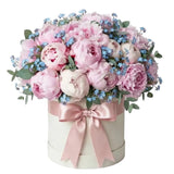 A round box bouquet filled with pink and white peonies, small blue flowers, and green eucalyptus leaves, tied with a pink satin bow on a white box against a white background.
